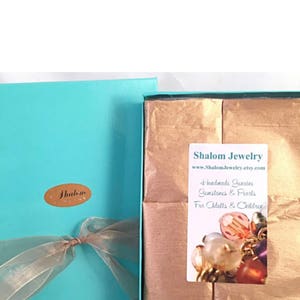 May include: A turquoise gift box with a gold ribbon and a label that says "Shalom Jewelry" is open to reveal brown tissue paper. The tissue paper has a label that says "Shalom Jewelry www.ShalomJewelry.etsy.com Handmade Genuine Gemstones & Pearls For Adults & Children".