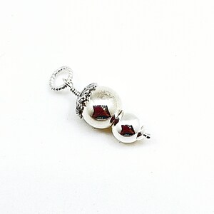 May include: A silver charm featuring two round beads, the top bead is larger and has a decorative design around the top, the bottom bead is smaller and smooth.