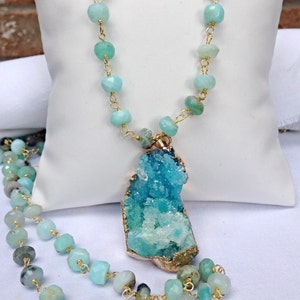May include: A gold chain necklace with a large, teal and green crystal pendant. The necklace is made of faceted, light blue beads.