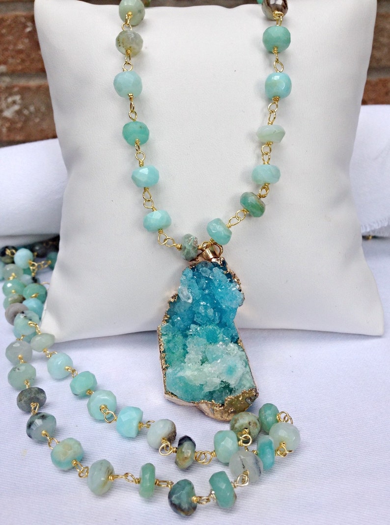 May include: A gold chain necklace with a large, teal and green crystal pendant. The necklace is made of faceted, light blue beads.