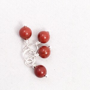 May include: Four red gemstone beads with silver wire loops. The beads are round and have a natural, marbled appearance.