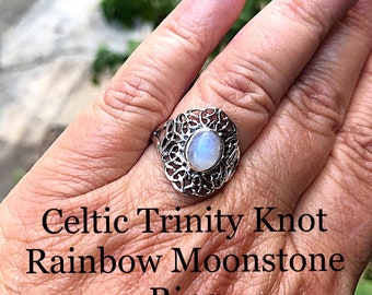 May include: A silver ring with a Celtic Trinity Knot design featuring a rainbow moonstone. The ring is on a person's finger.