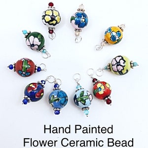 May include: Hand-painted ceramic bead earring charms with floral designs in various colors, including blue, yellow, red, and green. Each charm is a round bead with a unique flower pattern and silver-tone accents. The text reads: Hand Painted Flower Ceramic Bead Earring Charms.