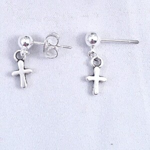 May include: A pair of silver-colored cross stud earrings. Each earring features a small cross charm dangling from a post with a round ball backing. The earrings are displayed on a white surface.