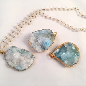May include: A gold chain necklace with white beads and three blue and white crystal pendants. The pendants are irregular in shape and have a gold trim.