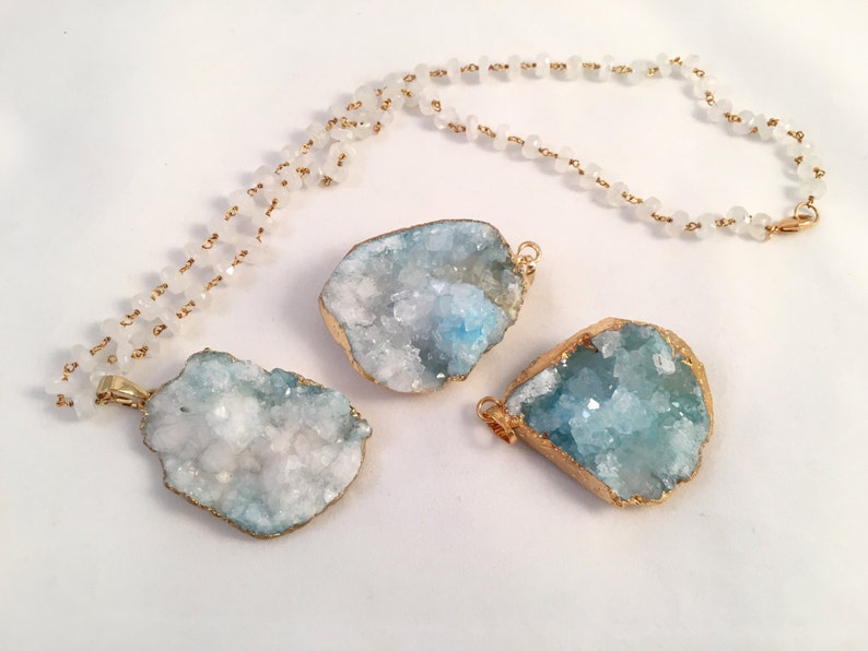 May include: A gold chain necklace with white beads and three blue and white crystal pendants. The pendants are irregular in shape and have a gold trim.