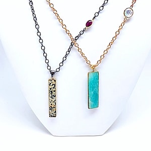 May include: Three necklaces with different pendants. The first necklace has a black chain and a rectangular pendant with a black and white speckled stone. The second necklace has a gold chain and a rectangular pendant with a turquoise stone. The third necklace has a gold chain with a white stone and a red stone.