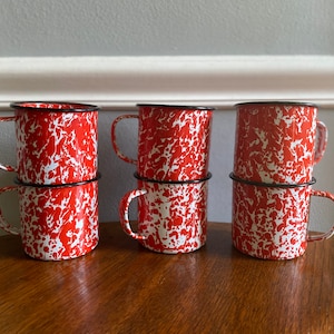 May include: Six red and white enamel mugs with black rims. The mugs are stacked in pairs and have a speckled pattern.