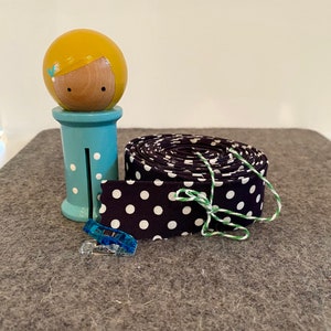 May include: A roll of black and white polka dot fabric with a green and white twine tied around it. A blue wooden doll with yellow hair is in the background.