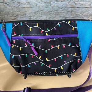 May include: A black fanny pack with a pattern of colorful string lights. The bag has a purple zipper and pull, blue side panels, and a purple adjustable strap. The bag is designed to be worn around the waist or across the body.
