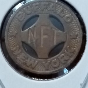 May include: A bronze-colored token with the text "BUFFALO NEW YORK" encircling a central design. The design features the letters "NFT" within a cross-shaped pattern. The token is encased in a clear plastic holder.