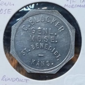 以下が含まれることがあります： 「C. BLOCKER GENL. M.D.S.E. ST. BENEDICT. KANS.」と書かれた銀色の八角形のトークン。トークンは保護ホルダーに入っており、手書きのメモが見えます。