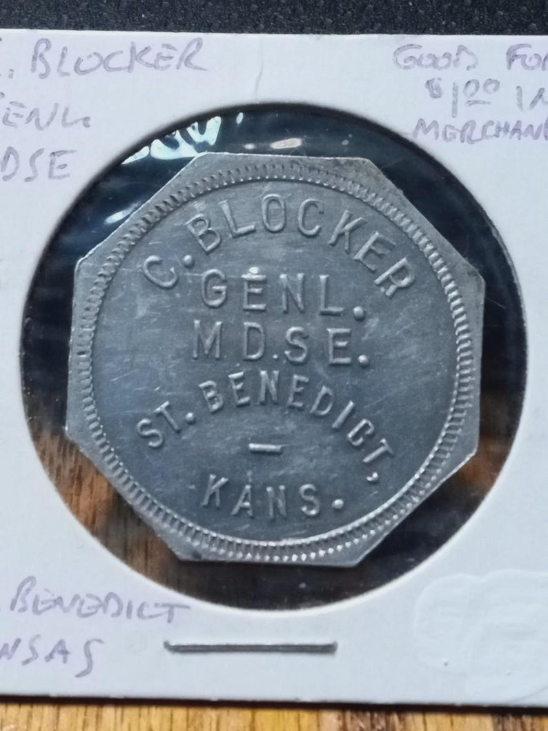 以下が含まれることがあります： 「C. BLOCKER GENL. M.D.S.E. ST. BENEDICT. KANS.」と書かれた銀色の八角形のトークン。トークンは保護ホルダーに入っており、手書きのメモが見えます。
