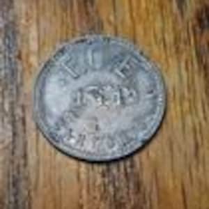 May include: A circular silver coin with the inscription "ICE" above the date "1873" and other illegible text. The coin shows signs of wear and is placed on a wooden surface.