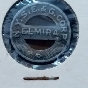 Puede incluir: Un objeto metálico circular plateado con el texto "S.E. & G. CORP" y "ELMIRA" en relieve. El objeto está encerrado en plástico transparente y colocado sobre un fondo blanco.