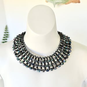 May include: A black iridescent beaded choker necklace with a wide, layered design. The necklace is made of small, faceted beads that create a shimmering effect.