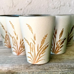 Può includere: Set di sei bicchieri in ceramica bianco sporco. Ogni bicchiere presenta un disegno di spighe di grano marrone. I bicchieri hanno una superficie leggermente strutturata e un bordo marrone scuro. Disposti a semicerchio.