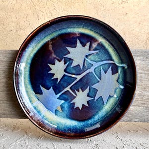 Puede incluir: Plato de cerámica hecho a mano con un esmalte azul oscuro y un diseño de estrella. El plato tiene un borde marrón y un borde azul claro y verde. El patrón de estrella está en un tono más claro de azul. Mide unos 25 cm de diámetro.