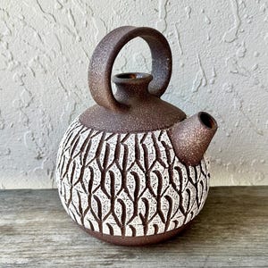 May include: A round ceramic teapot with a dark brown speckled finish. The upper portion and handle are smooth, while the lower body features a textured, white-filled geometric pattern of repeating vertical lines and carved shapes.