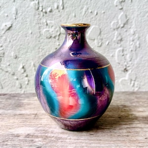 Pode incluir: Um pequeno vaso iridescente com corpo arredondado e pescoço estreito. O vaso apresenta uma mistura de cores, incluindo roxo, verde-azulado e vermelho, com detalhes dourados ao redor da borda e da base. O vaso é exibido em uma superfície de madeira.