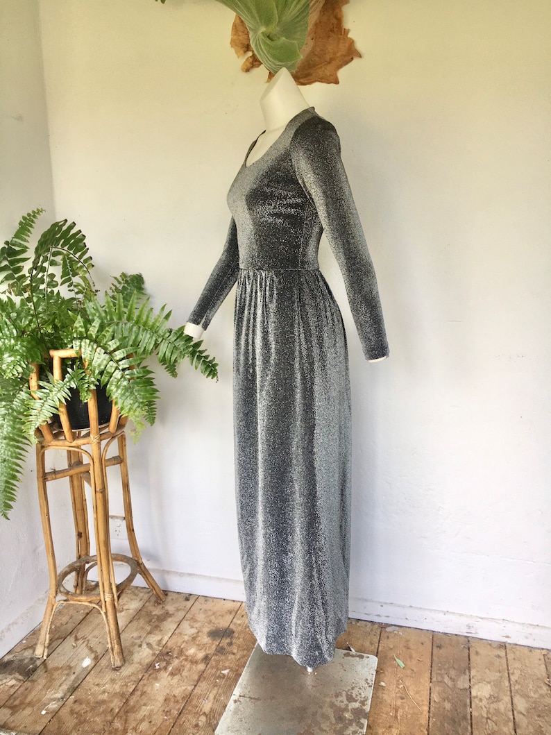 May include: A silver long-sleeved maxi dress with a sparkly texture. The dress has a scoop neckline and a fitted bodice. The skirt is flowing and has a slight A-line shape.