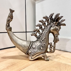 Pode incluir: Um recipiente de metal prateado em forma de criatura mítica com um corpo curvo semelhante a um chifre. A figura apresenta relevos intrincados, uma cabeça estilizada com crina e um pequeno animal pousado na ponta da cauda curva.