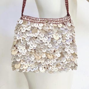 May include: A handbag covered in overlapping, circular, off-white and beige shell-like buttons. The bag features a dark reddish-brown crocheted wire top edge and a matching thin wire shoulder strap, creating a textured, layered appearance.