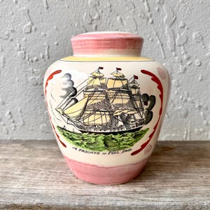 May include: A vintage ceramic vase with a ship illustration. The vase has a cream body with pink rim and base. The ship is in black and yellow, with the text "A Frigate in Full Sail" below. The vase is approximately 15 cm tall.