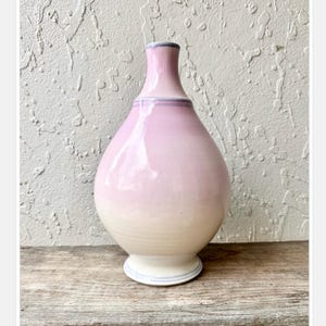 Pode incluir: Um vaso de cerâmica artesanal com um corpo bulboso e um pescoço fino. O vaso apresenta um gradiente de cores, passando de uma base branco creme a um tom rosa suave no topo. Uma faixa azul clara acentua a base e o pescoço.