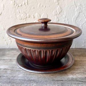 May include: A brown ceramic bowl with a lid and a matching saucer. The bowl has a ridged design around the sides and a small knob on top of the lid. The set is a warm brown color.
