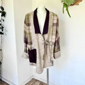 Op de afbeelding: Een beige, bruin en groen geruite jas met een donkerpaarse kraag en zak. De jas heeft een striksluiting en wordt op een mannequin getoond. De mouwen zijn omgeslagen en de jas lijkt van een wolachtig materiaal te zijn.