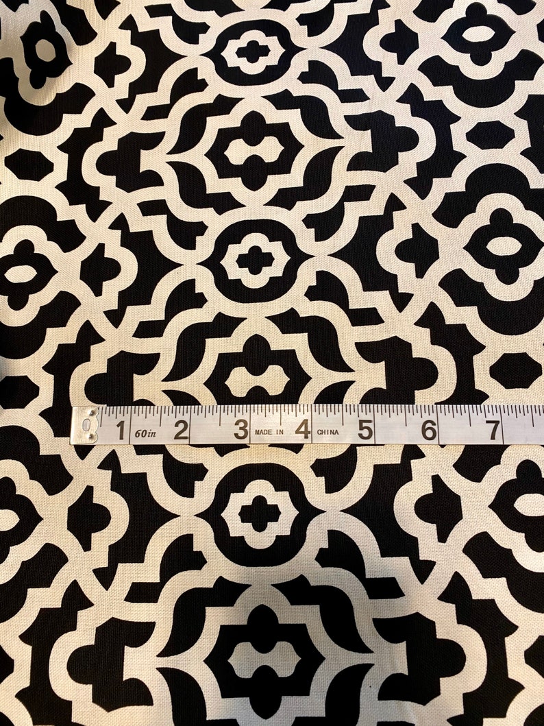 May include: Black and white fabric with a geometric pattern. The pattern is made up of repeating shapes that resemble a trellis or lattice. A measuring tape is placed on the fabric, showing that it is 60 inches wide.