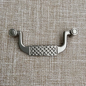May include: A silver cabinet pull with a decorative, woven design. The pull has two round knobs on either end.