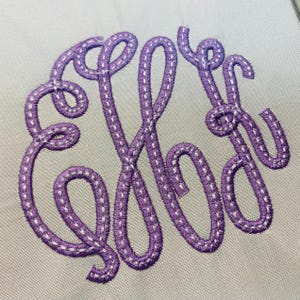 Könnte beinhalten: Eine hellgraue Stoffoberfläche mit einem großen, dekorativen Monogramm, das mit violettem Garn gestickt ist. Das Design besteht aus drei ineinander verschlungenen Buchstaben in Schreibschrift mit einem sichtbaren weißen Stichmuster.