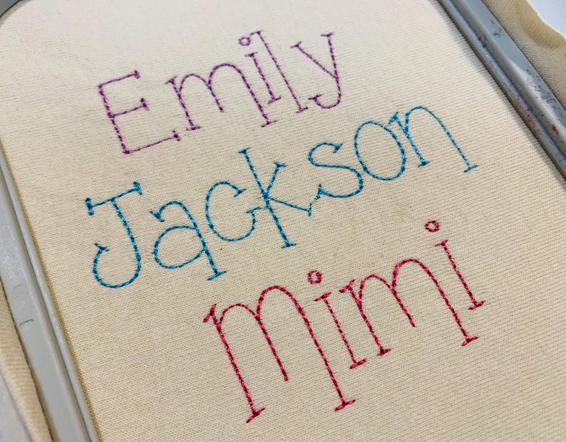 May include: An embroidery hoop containing off-white fabric with the name Emily stitched in purple, the name Jackson stitched in blue, and the name Mimi stitched in red. The lettering features a simple outline style with visible stitch details.