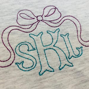 May include: A light grey heathered fabric featuring an embroidered design. The embroidery consists of a purple glittery bow positioned above the teal glittery letters S, K, and L, which are stitched in a decorative serif font style.