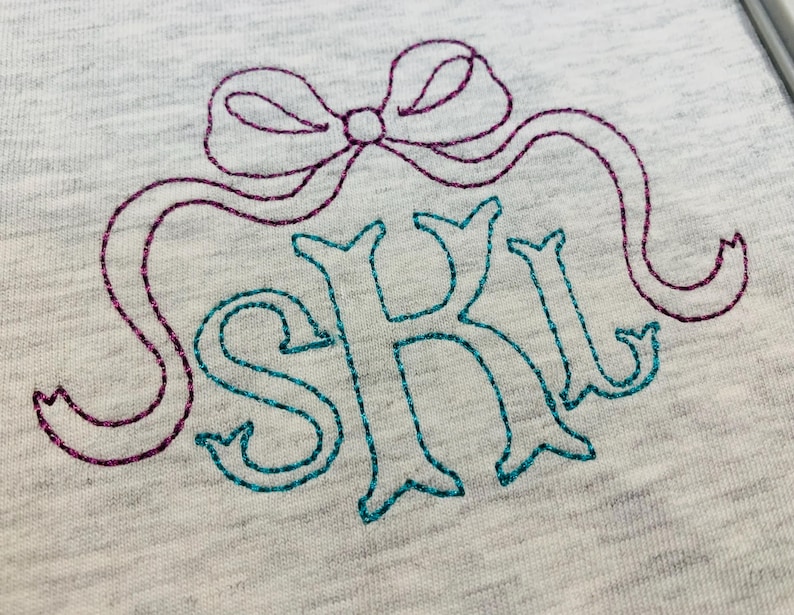 May include: A light grey heathered fabric featuring an embroidered design. The embroidery consists of a purple glittery bow positioned above the teal glittery letters S, K, and L, which are stitched in a decorative serif font style.