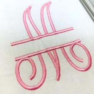 May include: A white fabric surface featuring a pink embroidered monogram design. The embroidery consists of a stylized letter M with a horizontal line separating the top and bottom sections, creating a split-letter effect with curved ends.