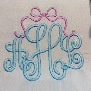 May include: A light blue embroidered monogram featuring the letters A, H, and L in a cursive script font, topped with a pink embroidered bow. The design is stitched onto a light grey fabric background, showcasing detailed thread work.