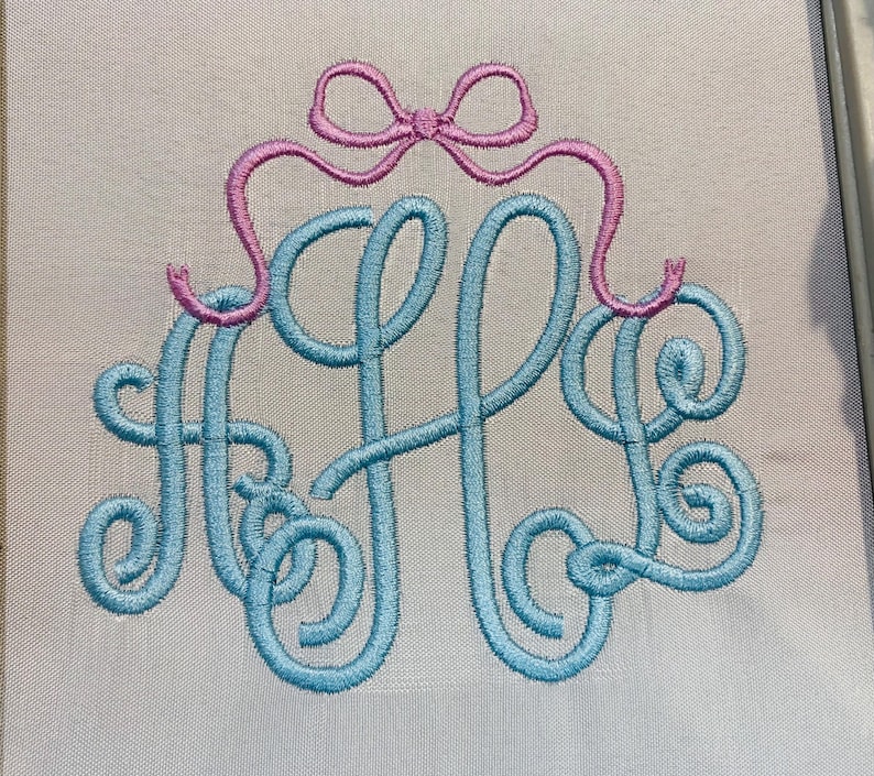 May include: A light blue embroidered monogram featuring the letters A, H, and L in a cursive script font, topped with a pink embroidered bow. The design is stitched onto a light grey fabric background, showcasing detailed thread work.