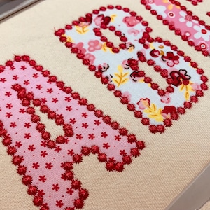 May include: A beige fabric surface features the letters A and B appliqu&eacute;d in floral patterned fabric. The letters are outlined with a dense, textured red thread embroidery stitch. The fabric patterns include pink and white floral motifs.