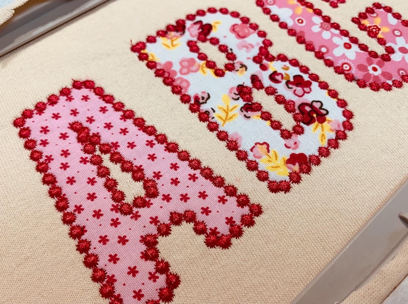 May include: A beige fabric surface features the letters A and B appliqu&eacute;d in floral patterned fabric. The letters are outlined with a dense, textured red thread embroidery stitch. The fabric patterns include pink and white floral motifs.