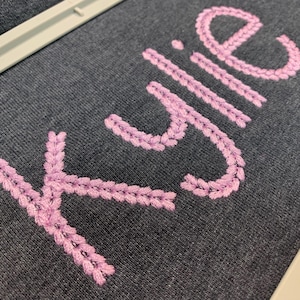May include: Close-up of a dark gray fabric with the name "Kylie" embroidered in light purple thread. The embroidery has a textured, woven appearance. The fabric is stretched within a white embroidery hoop.