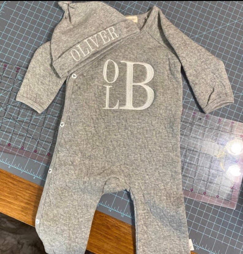 Op de afbeelding: Een grijze babyromper en bijpassende muts. De romper heeft een groot geborduurd "OLB" en drukknoopsluitingen. De muts is geborduurd met de naam "OLIVER". De set is gemaakt van een zachte, getextureerde stof.