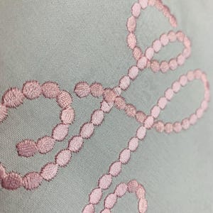 Op de afbeelding: Een close-up van een lichtblauwe stof met een roze geborduurd ontwerp van een reeks verbonden ovalen.