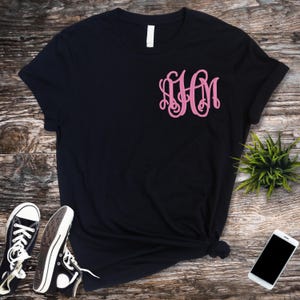 Op de afbeelding: Zwart t-shirt met een roze monogramontwerp op de voorkant. De afbeelding bevat zwarte canvas sneakers en een smartphone. Er is ook een kleine groene plant zichtbaar.