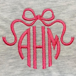May include: A light grey heathered fabric featuring a bright pink embroidered monogram with the letters AHM in a stylised font. Above the monogram is an embroidered pink bow with two long, curved ribbons extending outwards on both sides.