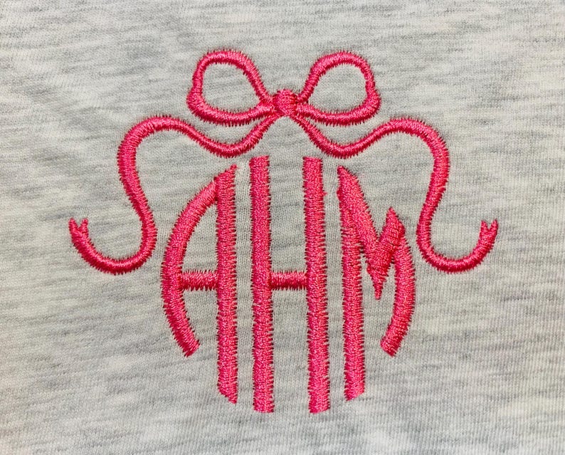 May include: A light grey heathered fabric featuring a bright pink embroidered monogram with the letters AHM in a stylised font. Above the monogram is an embroidered pink bow with two long, curved ribbons extending outwards on both sides.