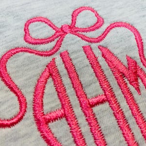 May include: Close-up of a light gray fabric with a bright pink embroidered monogram. The monogram features the letters "SAH" in a stylized script, topped with a bow design. The embroidery thread has a textured appearance.