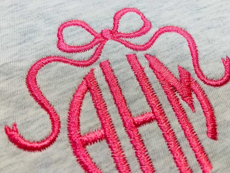 May include: Close-up of a light gray fabric with a bright pink embroidered monogram. The monogram features the letters "SAH" in a stylized script, topped with a bow design. The embroidery thread has a textured appearance.